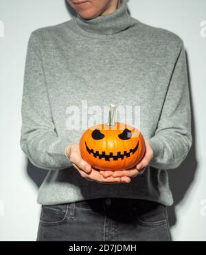 Crop femme méconnaissable tenue halloween face effrayante citrouille orange vif. Jeune femme en chandail en cachemire gris et Jean noir avec citrouille en forme de citrouille en forme de citrouille. Style d'instantané... Copier l'espace Banque D'Images