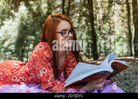 Souriant jeune belle femme à tête rouge lisant le livre tout en étant couché couverture de pique-nique avec journal au parc Banque D'Images