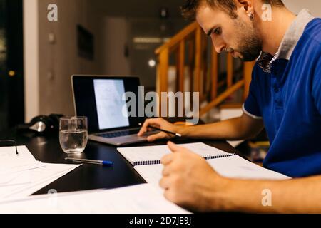 Un jeune homme s'est concentré sur ses devoirs tout en étant assis avec un ordinateur portable à la maison Banque D'Images