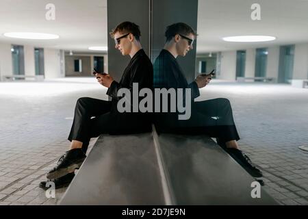 TRANS jeune homme portant des lunettes de soleil noires à l'aide d'un téléphone portable pendant assis au sous-sol Banque D'Images