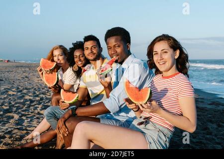 Des amis souriants qui mangent de la pastèque tout en étant assis sur la plage sous le soleil jour Banque D'Images