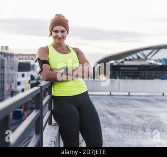 Jeune femme avec les bras croisés s'inclinant sur la main courante contre le ciel au coucher du soleil Banque D'Images