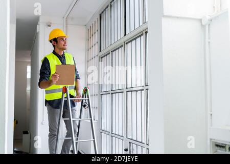 Homme portant un casque de travail et une veste debout sur une échelle fenêtre sur le chantier Banque D'Images