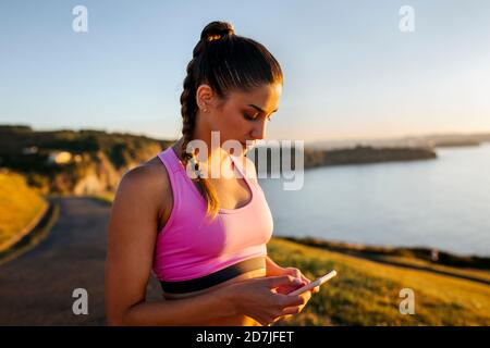 Jeune femme utilisant un téléphone intelligent en se tenant debout sur la falaise contre ciel clair au coucher du soleil Banque D'Images