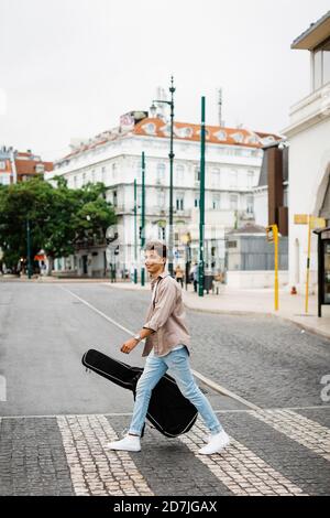 Homme souriant transportant de la guitare en traversant la rue en ville Banque D'Images