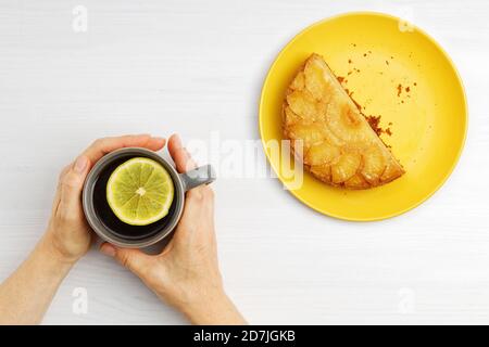 Les mains des femmes tiennent une tasse de thé chaude avec une tranche de citron et un morceau d'ananas à l'envers du gâteau. Vue de dessus. Banque D'Images