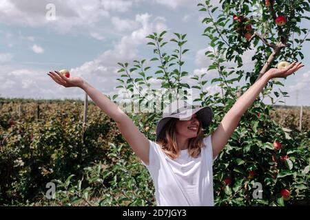 Femme insouciante portant un chapeau avec les bras levés tenant des pommes pendant debout dans un verger Banque D'Images