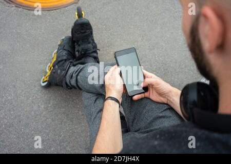 Gros plan d'un jeune homme portant des patins en ligne à l'aide d'un smartphone assis dans la rue Banque D'Images