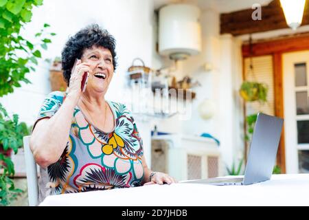 Femme âgée et gaie qui parle au téléphone intelligent tout en étant assise ordinateur portable dans la cour Banque D'Images