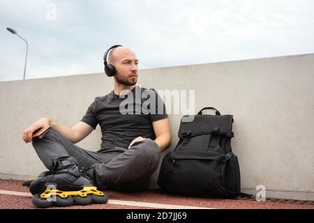 Jeune homme attentionné portant des patins en ligne qui écoutent de la musique tout en étant assis sur le pont Banque D'Images