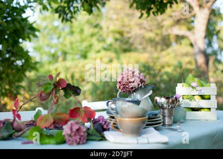 Vaisselle, abat-jour de lampe et carafe avec hortensias en fleur sur table basse dans le jardin Banque D'Images