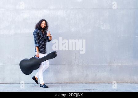 Homme souriant avec guitare montrant des coups de pied vers le haut tout en marchant contre mur gris Banque D'Images