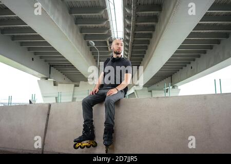 Jeune homme attentionné portant des patins en ligne tout en restant assis sur le maintien mur sous le pont Banque D'Images