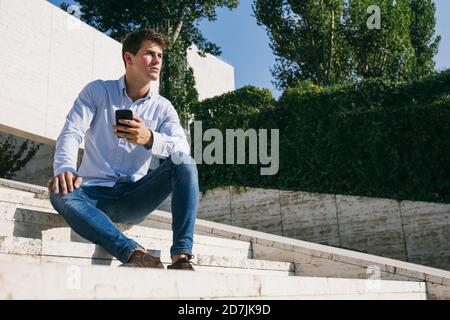 Un jeune homme charmant et attentionné assis avec un téléphone intelligent sur les marches contre les arbres en ville pendant la journée ensoleillée Banque D'Images