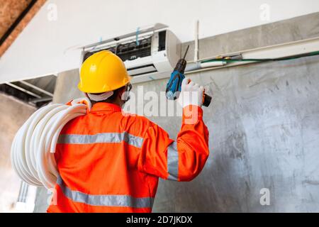 Technicien en climatisation mécanicien portant un masque et un casque pour prévenir la maladie, Covid 19 utilise actuellement une perceuse électrique pour installer le climatiseur. Banque D'Images