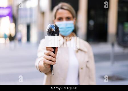 Femme journaliste portant un masque tenant un microphone en étant debout dans la rue en ville Banque D'Images