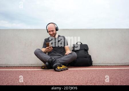 Jeune homme portant des patins en ligne à l'aide d'un smartphone assis avec sac à dos sur le pont Banque D'Images