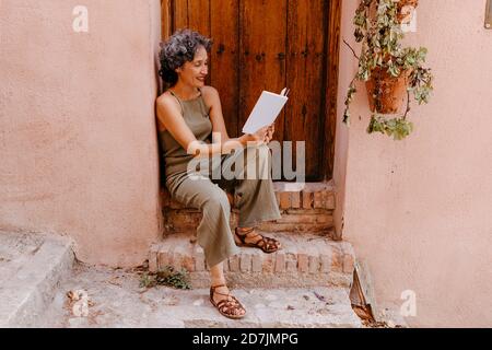 Femme mûre assise sur la porte tout en lisant le livre contre le bois porte Banque D'Images