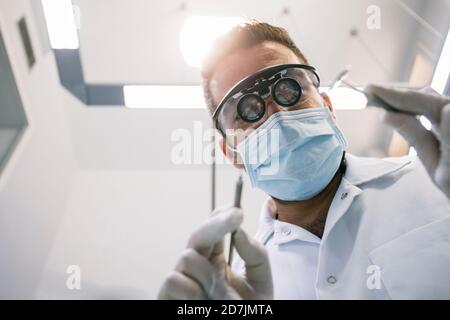 Dentiste de sexe masculin dans des loupes chirurgicales et un masque avec instruments médicaux pendant le traitement en clinique Banque D'Images