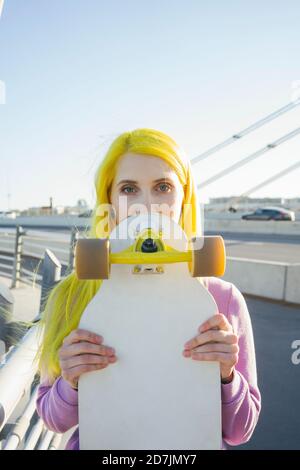 Jeune femme qui couvre le visage avec un skateboard en se tenant sur le pont par beau temps Banque D'Images