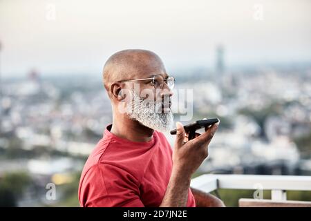 Homme chauve parlant à travers le smartphone tout en restant debout dans le bâtiment terrasse au coucher du soleil Banque D'Images