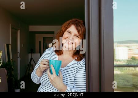 Femme souriante buvant du café tout en se tenant près de la fenêtre à la maison Banque D'Images
