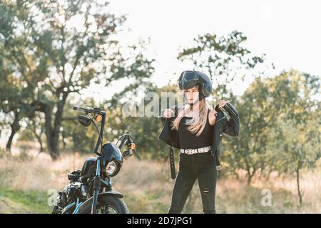 Blonde jeune femme motard vêtu d'une veste en cuir tout en restant debout moto Banque D'Images