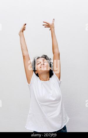 Femme mature debout avec les yeux fermés et les bras levés contre mur blanc Banque D'Images