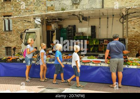 Une famille de touristes achetant dans une cabine de fruits et légumes au marché local de Piazza delle Erbe dans la vieille ville de San Gimignano, Toscane, Italie Banque D'Images