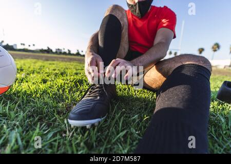 Footballeur dans un masque assis sur la pelouse et ficelant lacets Banque D'Images