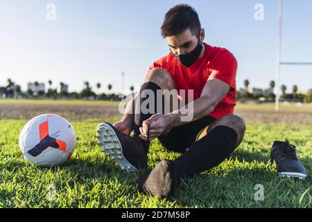 Footballeur dans un masque assis sur la pelouse et ficelant lacets Banque D'Images