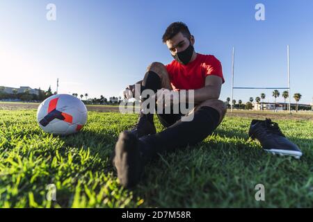 Footballeur dans un masque assis sur la pelouse et ficelant lacets Banque D'Images