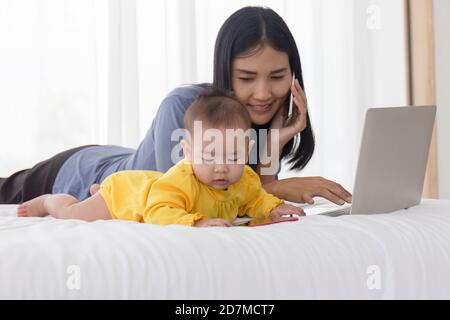 Un bébé asiatique joue à côté de sa mère dans le lit, avec sa mère parlant sur le téléphone cellulaire. Banque D'Images