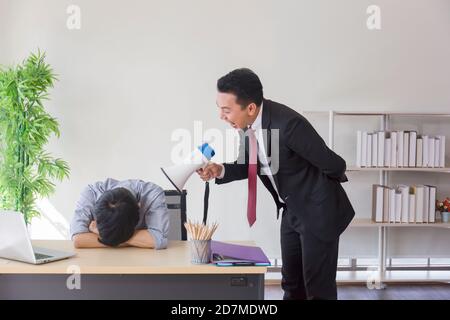 Un superviseur masculin asiatique utilise un mégaphone pour crier à ses subordonnés dormant sur une table de bureau. Banque D'Images