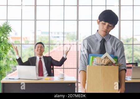 Un homme asiatique debout, malheureusement, tenant une boîte en carton contenant ses effets personnels et son Manager souriant derrière lui sur un bureau dans un bureau. Banque D'Images