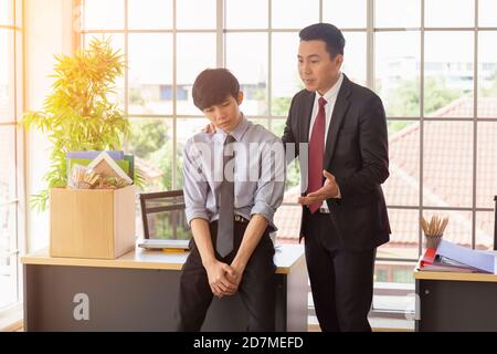 Le superviseur encourage un triste employé asiatique masculin à côté d'un bureau dans un bureau. Banque D'Images