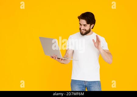 Joyeux homme barbu sur fond jaune parlant sur un lien vidéo . un jeune homme dans un t-shirt blanc avec un ordinateur portable dans ses mains regarde l'écran Banque D'Images
