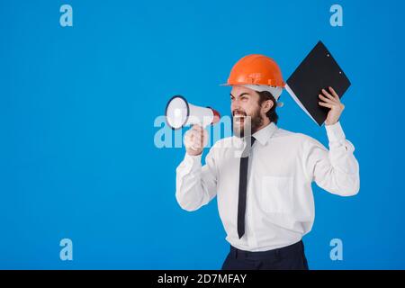 Un jeune homme barbu de construction dans une chemise blanche et un casque de construction sur fond bleu. Un ingénieur-architecte industriel crie Banque D'Images