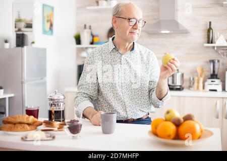 Homme mature tenant une pomme verte avant de prendre un morsure pendant le petit déjeuner dans la cuisine. Personne âgée à la retraite mangeant de la pomme pendant le petit-déjeuner dans une cuisine moderne et confortable. Sain vert heureux style de vie frais Banque D'Images