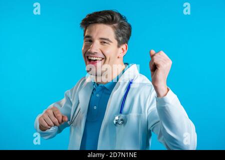 Sourire médecin danse drôle, succès et chance au travail. Jeune homme de doc beau en professionnel médical blanc manteau est isolé sur le studio bleu Banque D'Images