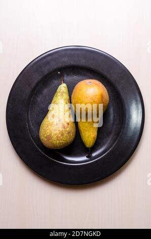 Deux fruits à la poire, poires, Pyrus, plat, posé sur une plaque ronde noire sombre sur une table ou un bureau en bois, photographie alimentaire, gros plan, espace de copie Banque D'Images