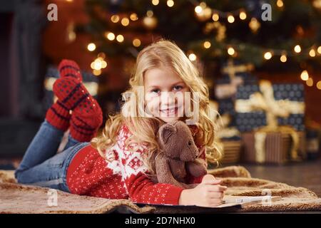 Adorable petite fille en chandail rouge festif couché avec ours en peluche à l'intérieur pour célébrer le nouvel an Banque D'Images