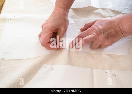 la main des tailleurs fixe les dispositions de coupe de papier de la robe à calico tissu avec épingles sur une table en bois à la maison Banque D'Images