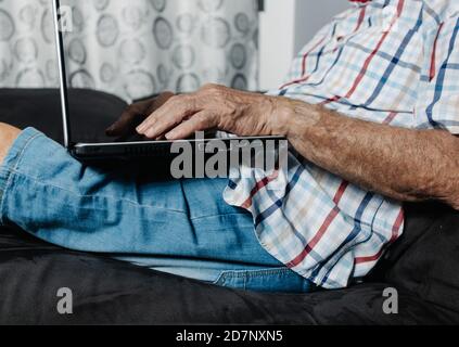 Vue latérale de la récolte homme âgé méconnu dans des vêtements décontractés assis sur un canapé et tapant sur un ordinateur portable tout en travaillant à distance de la maison. Banque D'Images