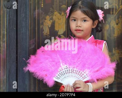 Habillée, jolie petite fille chinoise thaïlandaise pose avec un éventail de plumes à main rose, pendant le nouvel an chinois, à l'intérieur du Temple de Lotus Dragon. Banque D'Images