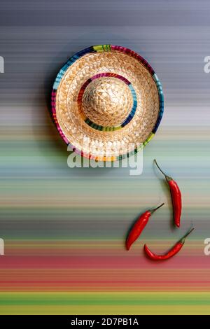Chapeau de paille mexicain coloré à côté de trois piments rouges chauds sur un fond à rayures colorées. Inspiration mexicaine avec l'espace de copie. Banque D'Images