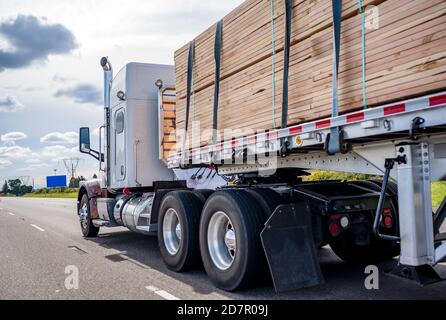 Blanc classique puissant capot américain grand rig semi-camion professionnel tracteur transportant une cargaison de bois de charpente serrée sur un semi-plateau exécution de la remorque Banque D'Images