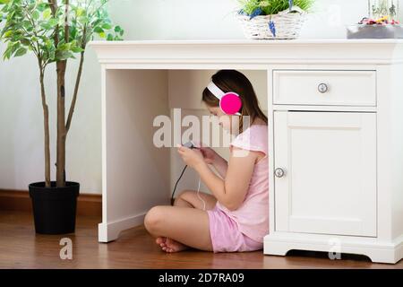 Enfant avec casque et téléphone portable sous le bureau. Apprentissage à distance en ligne dans des endroits inhabituels. Examen à distance pendant le verrouillage. Adolescent à l'écoute de musi Banque D'Images