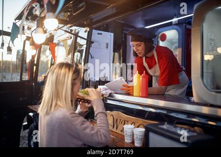 une jeune cliente caucasienne mange un sandwich devant le service de restauration rapide, en donnant des suggestions à un bel employé Banque D'Images