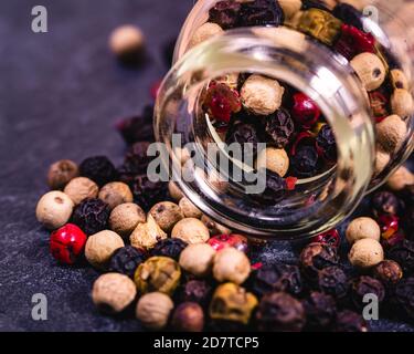 Gros plan de 4 types de grains de poivre qui débordent d'un bouteille en verre sur une surface texturée foncée Banque D'Images